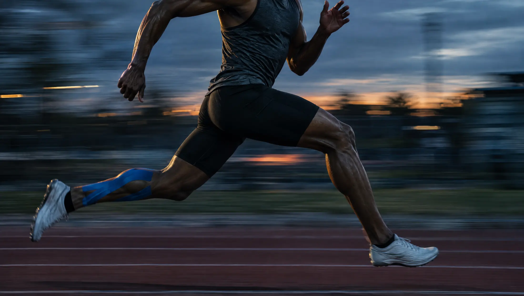 A sprinter with blue kinesiology tape on one calf, mid-stride on a track at dusk
